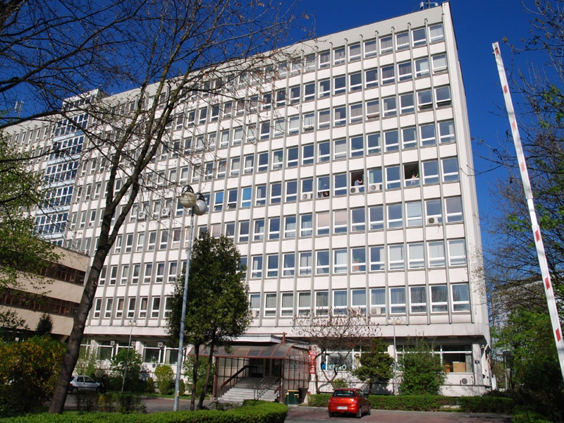 Zdjęcie przedstawia widok na modernistyczny biurowiec otoczony zielenią.